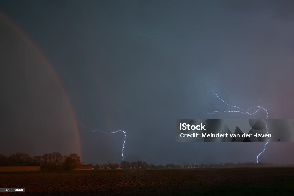 Lightning storm with rainbow Lightning bolts strike down to earth while the setting sun illuminates the plain landscape and creates a rainbow. Agricultural Field Stock Photo