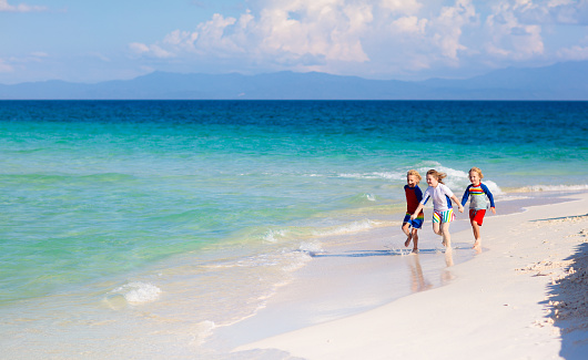 Kids playing on tropical beach. Children swim and play at sea on summer family vacation. Sand and water fun, sun protection for young child. Little boy and girl running and jumping at ocean shore.