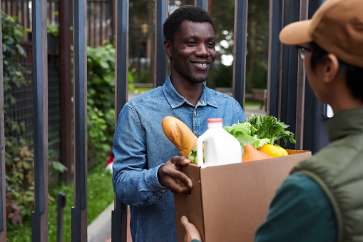 Portrait of smiling black man delivering groceries in city and holding box with fresh vegetables