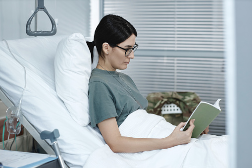 Female soldier in eyeglasses reading a book resting on bed in hospital ward during treatment