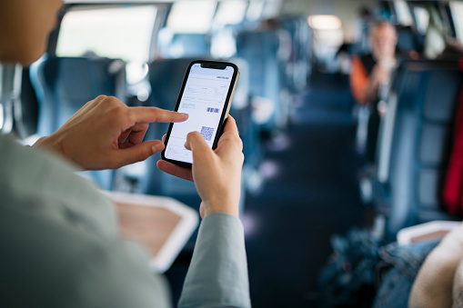 Woman looking at electronic train ticket on her phone screen while standing in train coach. Over the shoulder view of a female traveler in train looking at her seat number on train boarding pass over her mobile phone.