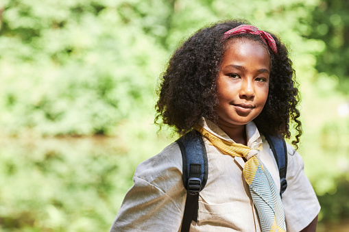 Waist up portrait of cute black girl as girl scout looking at camera outdoors in forest, copy space