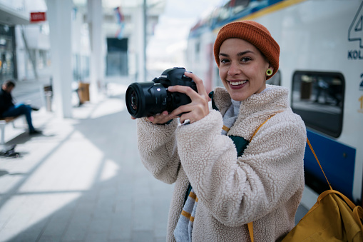 Smiling woman traveler taking a photos with professional camera on railway station. Caucasian female traveler photographing with her dslr camera on train platform.
