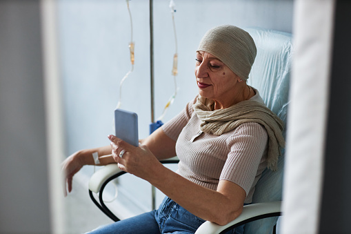 Side view portrait of senior woman sitting in comfortable chair with IV drip and using smartphone