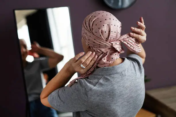 Back view of unrecognizable bald woman tying headscarf looking in mirror