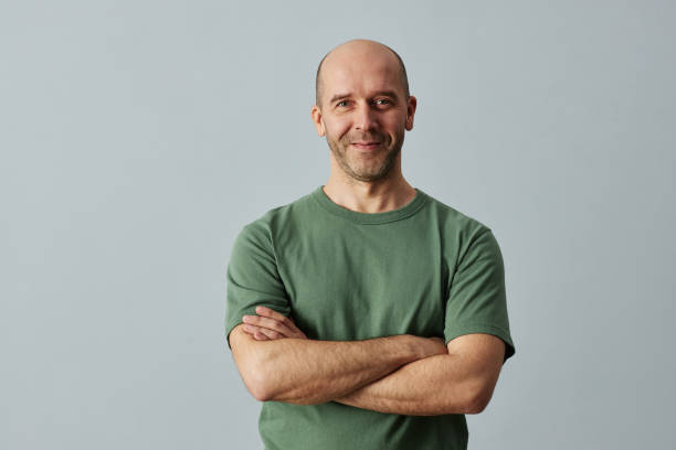 hombre calvo sonriendo a la cámara de pie con los brazos cruzados - portraite fotografías e imágenes de stock