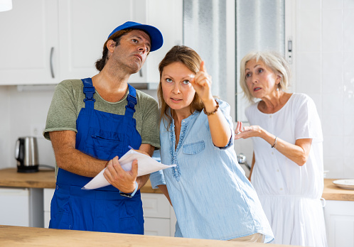 Professional male repairman in uniform taking notes in the presence of two women of different ages in the kitchen