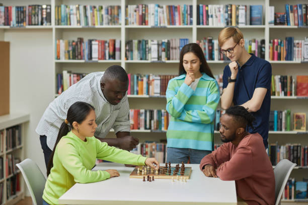istock Two young people playing chess in library with group of friends watching 1485548998