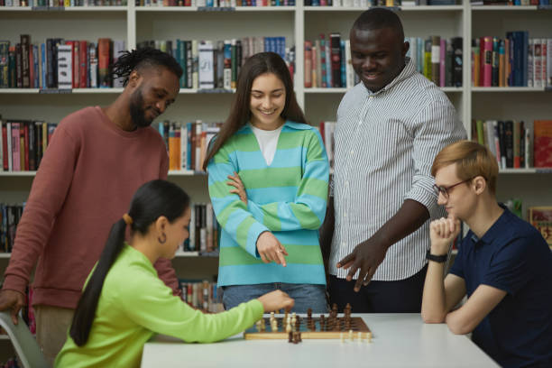 Two young people playing chess with group of friends watching and cheering Side view at two young people playing chess with diverse group of friends watching and cheering friends playing chess stock pictures, royalty-free photos & images