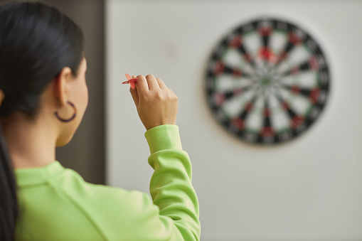 Back view closeup of young woman playing darts and aiming shot, copy space
