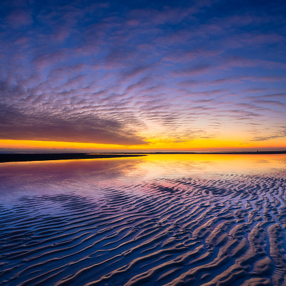 A beach during sunrise. The sky is beautifully colored in pink, purple and orange by the setting sun. There are some clouds in the sky and the sea is moving wildly.