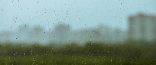 istock Atmospheric minimal backdrop with rain droplets on glass. Wet window with rainy drops and dirt spots closeup. Blurry buildings and green trees under blue sky against dirty window glass with raindrops. 1485574886