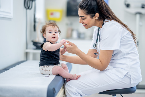 A sweet little baby boy sits up on an exam table with the assistance of a nurse.  He is dressed casually and appears happy as the nurse looks him over.