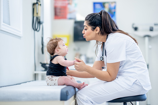 A sweet little baby boy sits up on an exam table with the assistance of a nurse.  He is dressed casually and appears happy as the nurse looks him over.