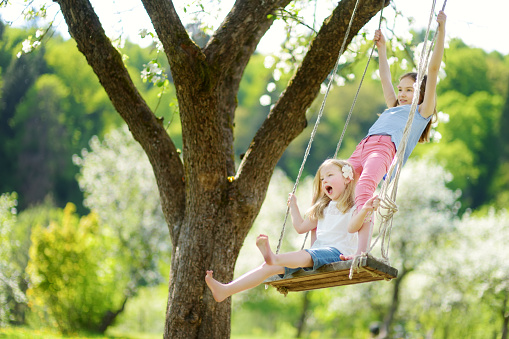 A single Mom pushes her daughter on a swing while spending some quality time together outside. The little girl is dressed casually and wearing a hat to keep the warm summer sun off her face. She is giggling with each push her mother gives her.