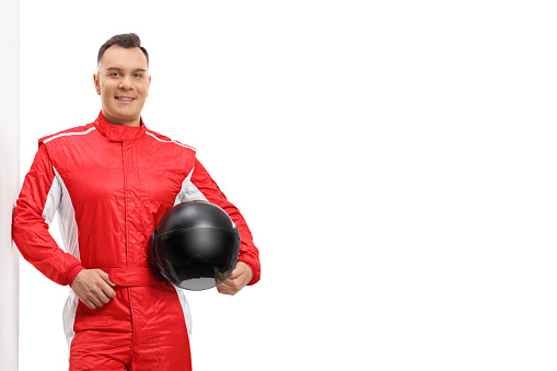 Racer in a red suit leaning on a wall and holding a black helmet isolated on white background