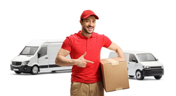 Delivery guy standing in front of transport vans and pointing to a box isolated on white background