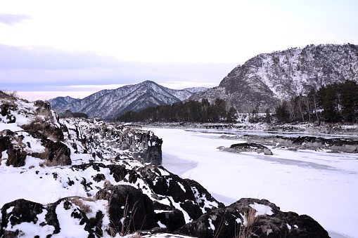 Rocky banks of a beautiful frozen river in a picturesque mountain valley covered with snow in the early winter morning. Katun river, Altai, Siberia, Russia.