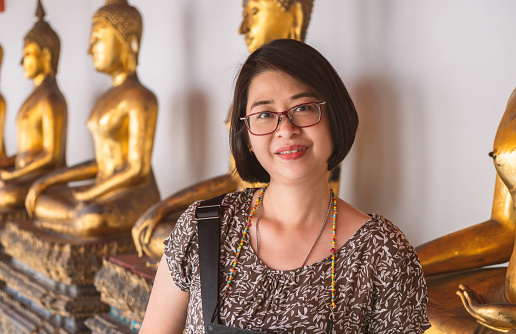 Portrait of an Asian tourist woman in a temple of Thailand, middle aged woman. wears eyeglasses, smiling and looking at camera, blurred background or a row golden Buddha, landscape image.