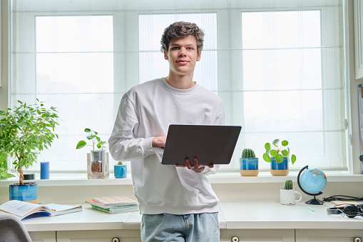 Portrait of friendly young college student guy with laptop at home. Handsome male 18-20 years old looking at camera. Online internet technologies, remote e-learning, study, leisure, communication
