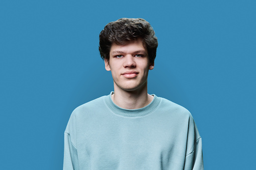 Headshot portrait of young male 18-20 years old in casual sweatshirt, on blue studio background. Handsome guy with curly hair looking at camera. Lifestyle, youth concept