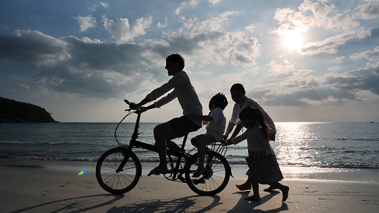 Asian family On Summer Vacation Four people Father, mother, daughter and son riding bicycles and walking In the evening the sunset on the beach. Travel and Relaxation