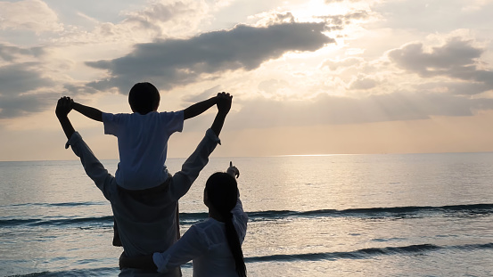 Happy Asian Family On Summer Vacation, a boy on the shoulders of a father open arms and looking at the sky on the beach Background sunset in the sea. Travel and Relaxation