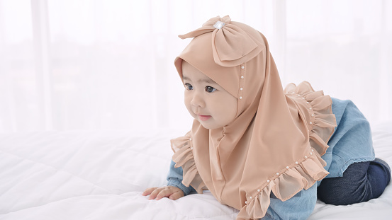 Muslim Asian, Little baby 6 months old in traditional hijab clothing crawling on the bed at home