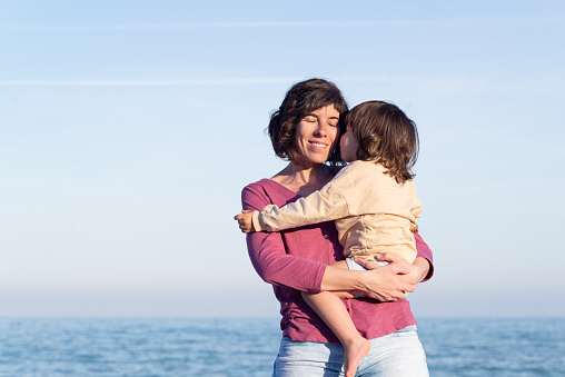 Smiling mother carrying her little daughter while she is kissing her with the sea on the background.