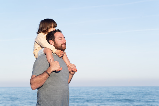 Relaxed man carrying her little daughter on shoulders and breathing fresh air with the sea on the background.