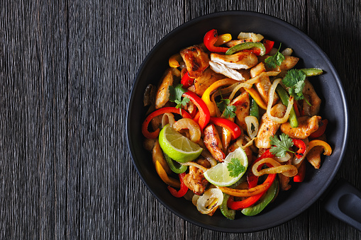 chicken fajitas of fried chicken breast strips, onion, julienned green, yellow and red bell peppers on skillet on dark wood table, horizontal view from above, flat lay, close-up, copy space