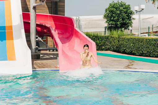 Young woman slide in pool after going down water slide at sunny day in water Aqua park. Refreshing at heat weather, active vacation and healthy lifestyle. Happy summer.