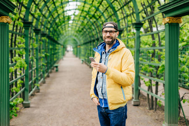 junger student mit bart mit stilvoller kleidung vor grünem hintergrund, der ein mobiltelefon zum herunterladen von videos verwendet. bärtiger hipster in gelbem anorak, der sich im freien ausruht und nachrichten tippt - dimple male human face men stock-fotos und bilder