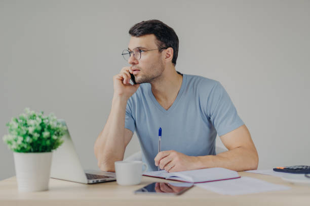 istock Serious male finance manager talks on mobile phone, solves problems with partner, writes some notes in diary, arrange meeting, surrounded with laptop computer, isolated over grey background. 1485673505