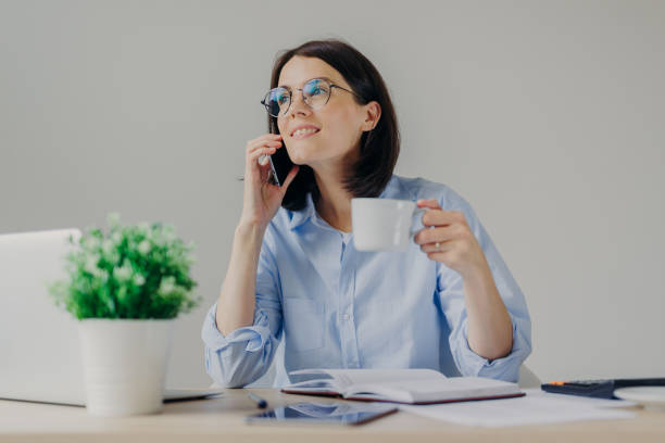 istock Gorgeous pleased businesswoman has cell phone conversation while has coffe break after working on notebook, shares her success with close person, isolated over grey background. Occupation concept 1485674438