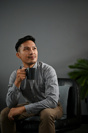 Portrait of determined and handsome millennial Asian man in casual clothes sits in a modern living room with his coffee cup.