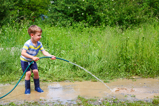 Boy playing with water hose. Making a big puddle of mudd. Standing in rubber boots.