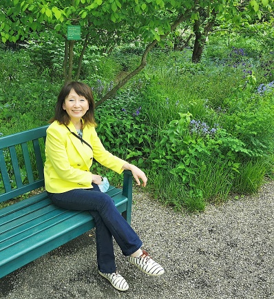 Happy smiling attractive middle aged brunette woman in yellow color blazer sits on bench in park.