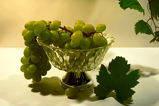 A bunch of white grapes in a glass bowl with a silver base. A grape vine and leaf serve as decoration