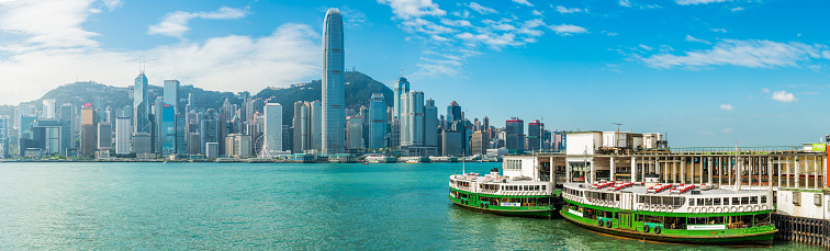 Panoramic view across the blue waters of Victoria Harbour and iconic Star Ferries overlooked by the crowded skyscraper cityscape of Hong Kong Island, China.
