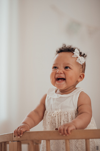Candid authentic portrait of cute african american diverse female child at home interior of earth colors. Happy swarthy infant baby stands in kids bed in white lace dress and bow headband at bedroom