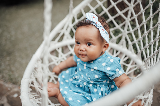 Swarthy little girl in blue polka dot dress has positive emotion sitting on white macrame cotton rope cocoon hanging. African american baby infant relaxes on swing cozy place on open air