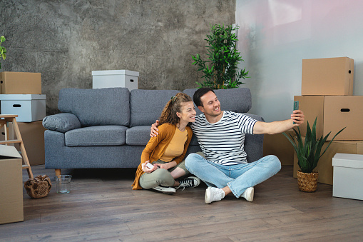 The young Caucasian couple, sitting on the floor in their new apartment and taking a selfie with their mobile phone