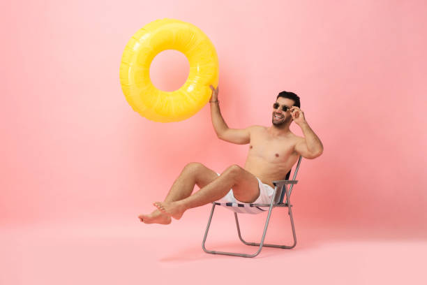 glücklicher hemdloser kaukasischer touristenmann, der auf einem strandkorb sitzt und einen schwimmring in isoliertem rosa farbstudiohintergrund hält, sommer-berufungskonzept - beach men isolated tourist stock-fotos und bilder