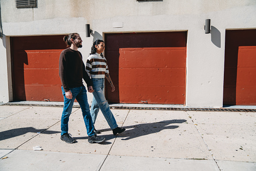 An hipster couple is walking together on a sidewalk in Bushwick. They are hanging out together.