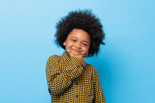 Cute African mixed race afro boy posing and smiling with hand on chin in blue color isolated background studio shot