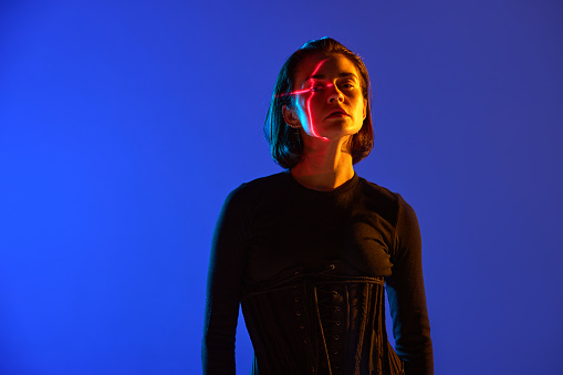 Portrait with attractive woman wearing black clothes and reflection red line on face and looking at camera over blue mode neon background. Concept of digital art, online, fashion, cyberpunk, futurism