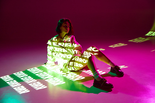 Error, mistake. Portrait of dreaming girl sitting on floor with digital neon filter lights with inscryption on body on pink mode background. Concept of digital art, fashion, cyberpunk, futurism