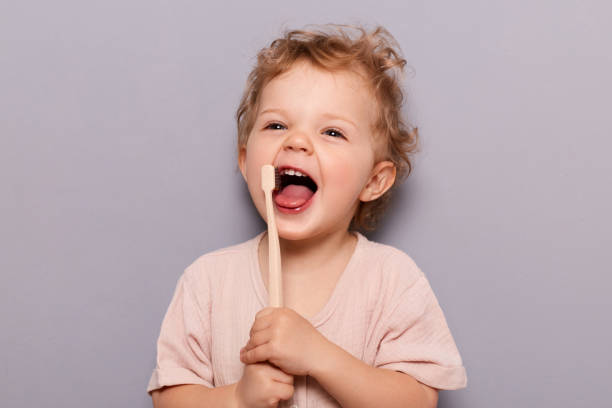 ritratto di bambina allegra che ride felice con i capelli biondi ricci che si lavano i denti, prendendosi cura dell'igiene dentale in piedi isolata su sfondo grigio. - healthy lifestyle toothbrush caucasian one person foto e immagini stock