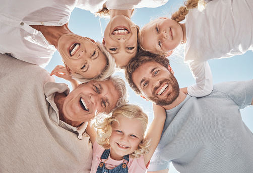 Happy family, huddle and smile below in trust, community or support together against a blue sky. Portrait of grandparents, parents and children hugging, smiling or bonding for holiday break in nature